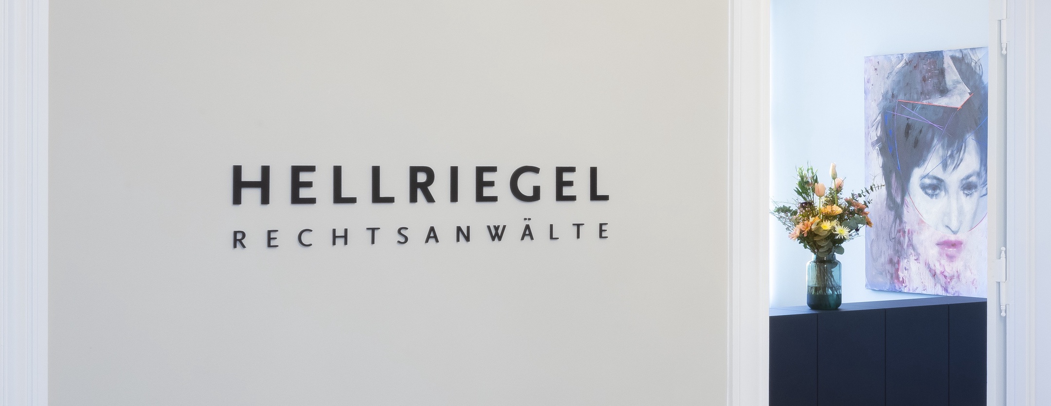 Eingangsbereich der Kanzlei HELLRIEGEL. Man sieht eine Wand auf der der Schriftzug "Hellriegel Rechtsanwälte" steht. Hinter der Wand guckt ein Tresen mit Blumen darauf hervor.