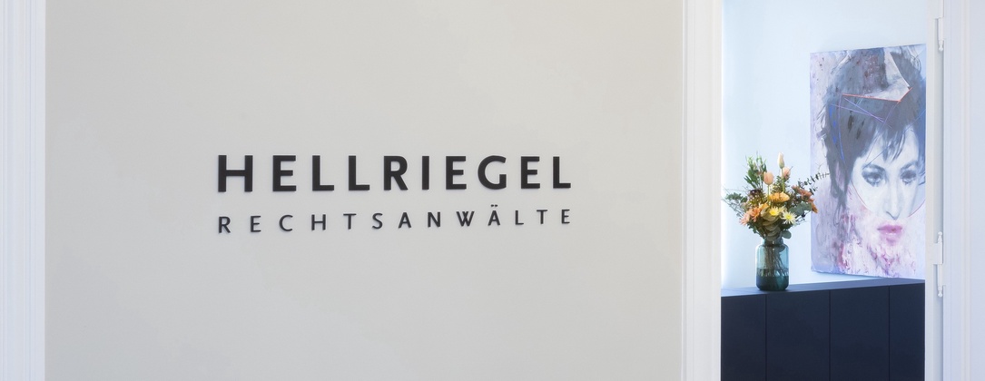 Eingangsbereich der Kanzlei HELLRIEGEL. Man sieht eine Wand auf der der Schriftzug "Hellriegel Rechtsanwälte" steht. Hinter der Wand guckt ein Tresen mit Blumen darauf hervor.