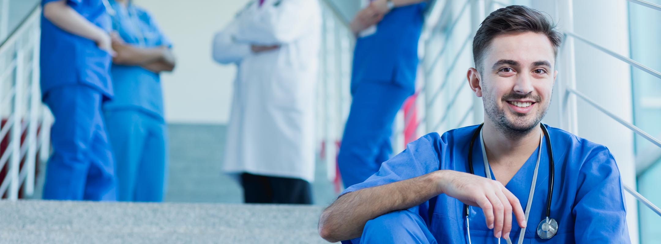 Ein männlicher Pfleger sitzt auf einer Treppe, im Hintergrund sieht man einen Arzt und weitere Pflegekräfte.