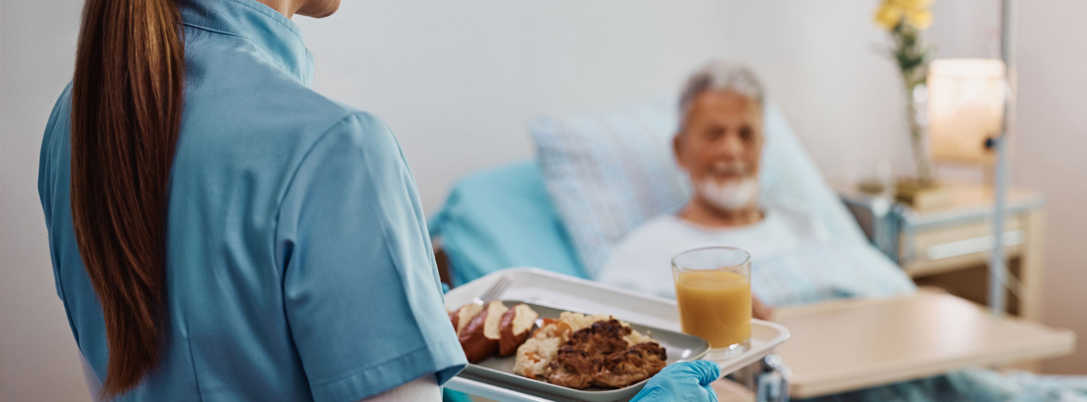 Eine Pflegerin bringt ein Tablett mit Essen an ein Krankenbett